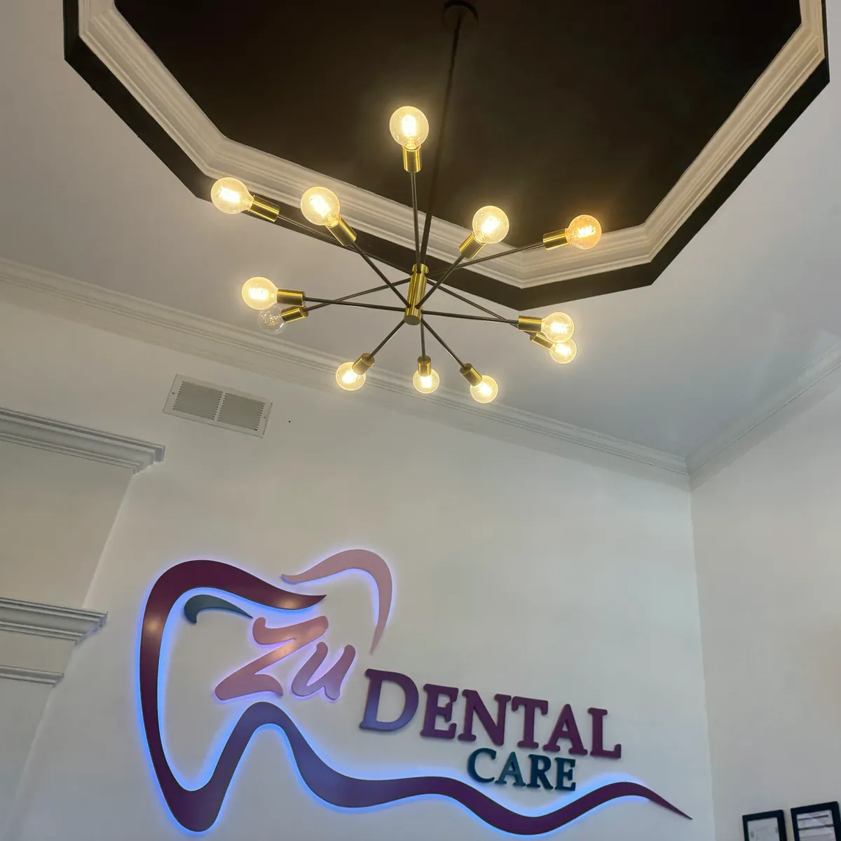 Una lámpara de bombillas doradas bajo el techo de la recepción de Zu Dental Care, con el logo iluminado debajo.